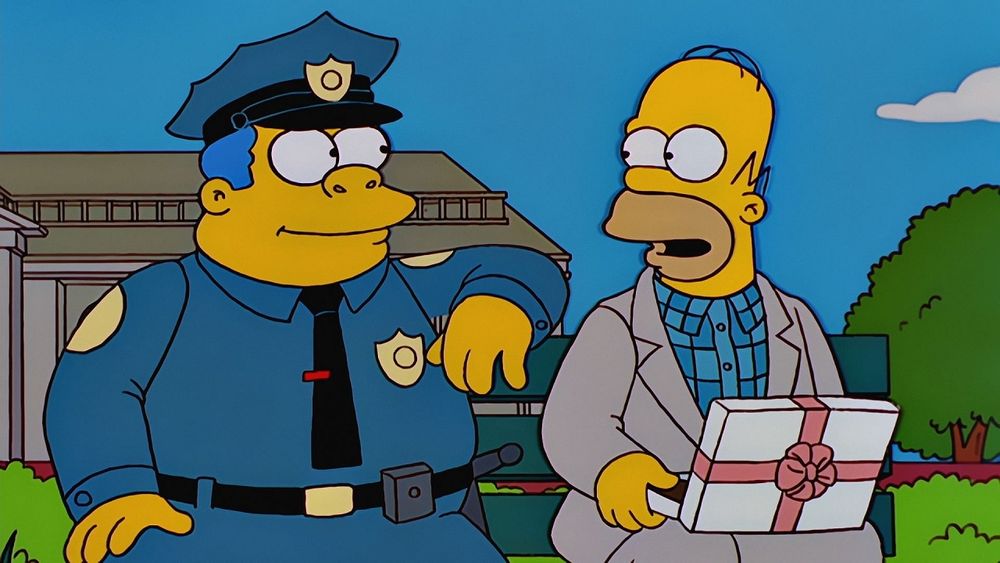 A screenshot from The Simpsons. It parodies the movie Forrest Gump. Chief Wiggum and Homer Simpson sit on a bench together. Wiggum is looking at Homer and has his arm resting on the top of the bench. Homer is looking back and holding a box of chocolates.