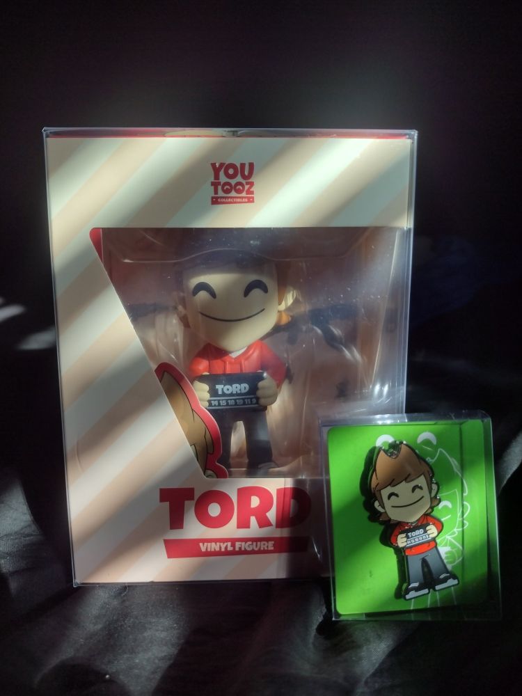A picture of the Tord Eddsworld Youtooz figure still in its box. Leaning against it is a pin of the figure