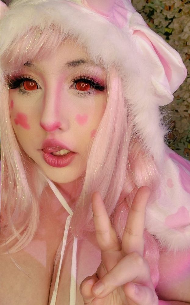 A photo of me  from the chest up, dressed as a pink spotted cow, i have big red eyes and long sparkley pink hair, im wearing a pink cow spotted hood with ears and horns and a pink spotted bikini with white fur trim. Im holding my hand up with a peace sign and have my lip poked to the side.