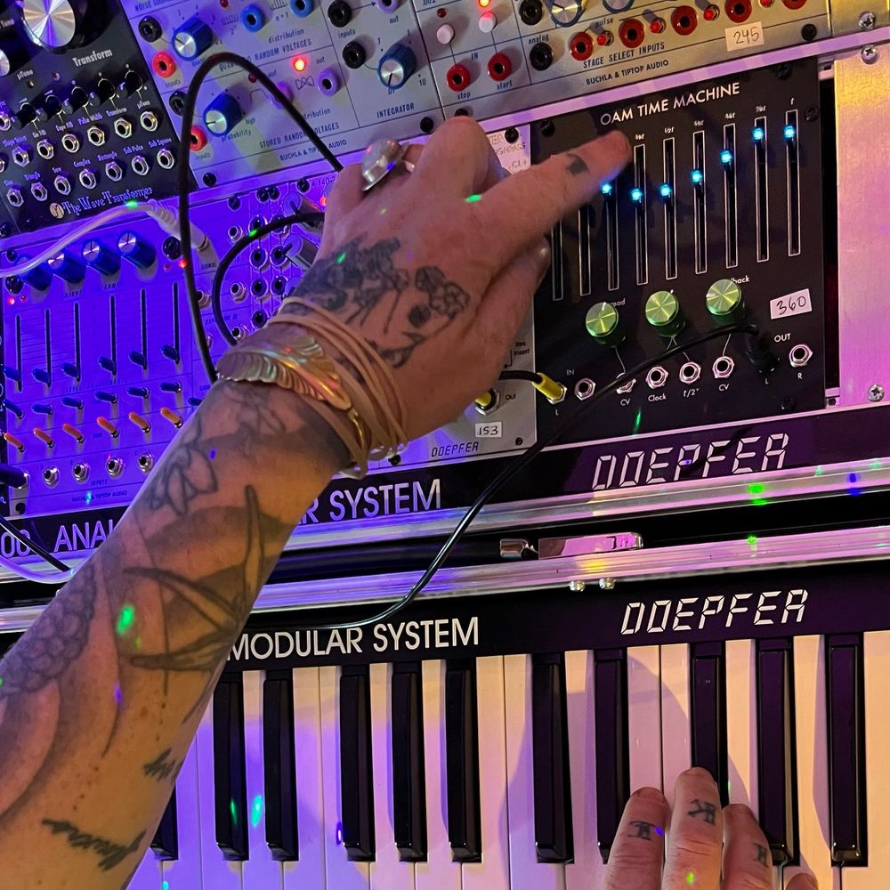 two beautifully inked up hands, reaching over a piano keyboard towards a large modular synth system. specifically, they are reaching to a large eurorack delay with blue LED sliders and green aluminum knobs.