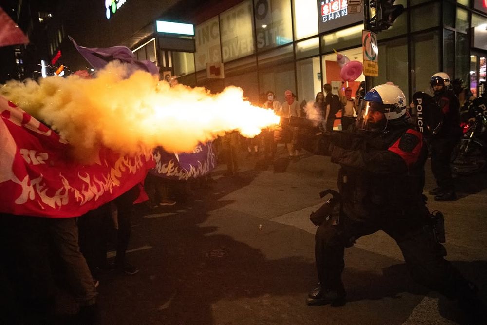 A man in riot gear with a helmet and red armband fires a projectile weapon in the direction of people holding a banner. Flame and smoke shoot out from the gun.