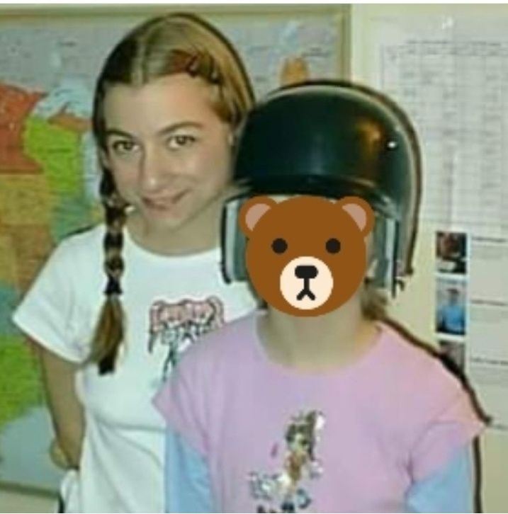 Me with my daughter when she was 7 years old. We just finished a softball game so she is still wearing her black helmet. She is also wearing a pink tee shirt with a cartoon girl on it. My daughter's face is hidden by a bear emoji for privacy. I am standing slightly behind her and have my hair in two braids. I am wearing a white tee shirt with a cartoon girl wearing sunglasses on it. I have long dark blond hair and brown eyes. AnUS map is on the wall behind us as are calendars.