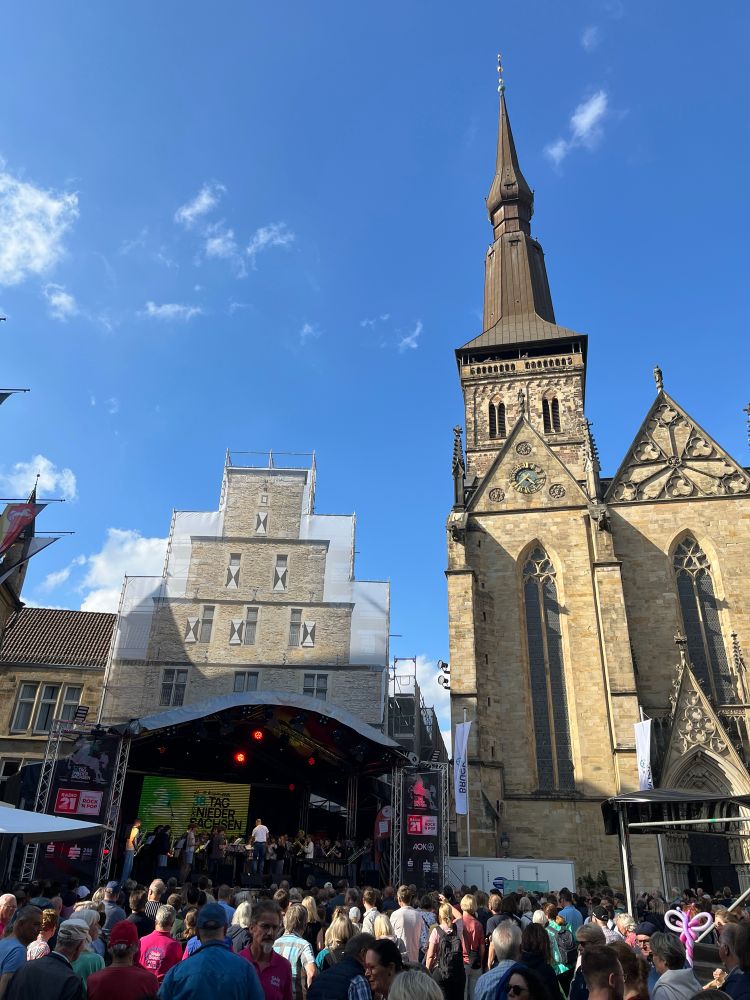 Blick auf den Marktplatz mit vielen Menschen vor einer Bühne. Links ein altes Steingebäude, rechts ein Kirchturm mit Kupferdach. Blauer Himmel