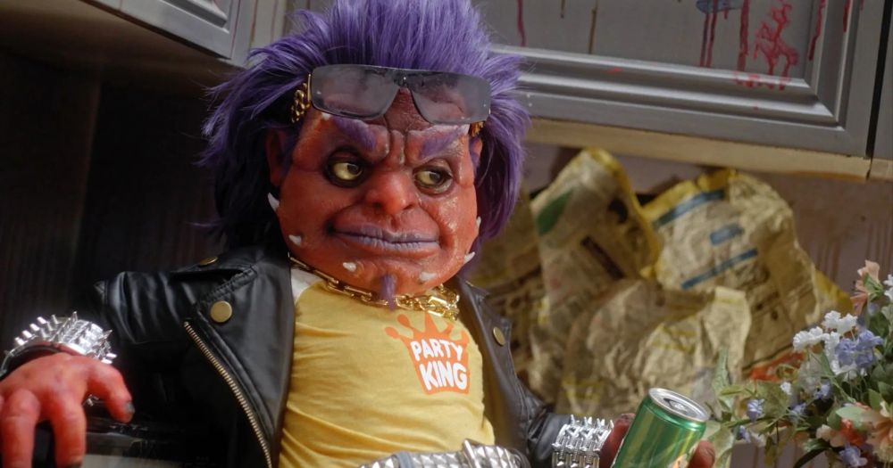 Frankie Freako, a puppet character with red demonic face, a yellow T-shirt saying "party king", purple hair, punk jewellery, a leather jacket, and a beer.