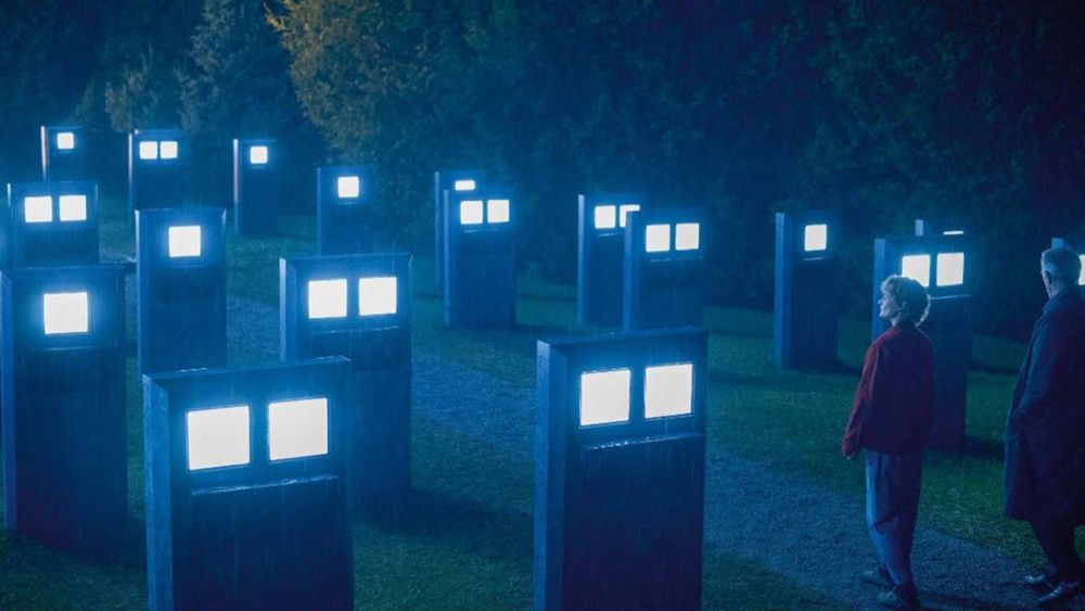 Still from The Shrouds - two people explore a graveyard at night, the gravestones all have brightly lit screens.