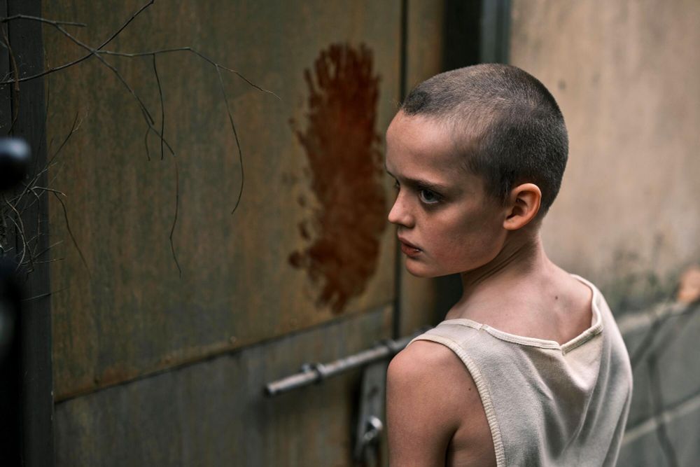 Sora Wong as Piper, a young girl with a shaved head, in Bring Her Back. There's a red stain on the grotty wall behind her.