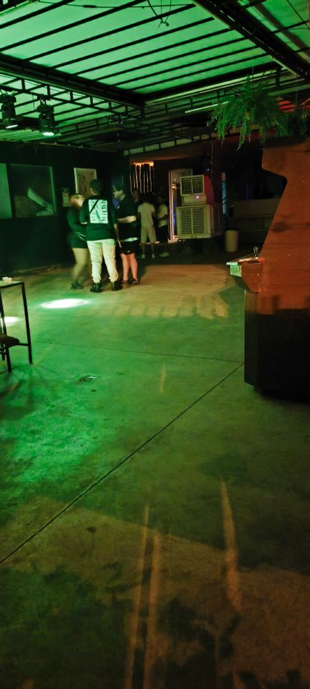 A imagem mostra um ambiente interno amplo com iluminação em tons de verde. O teto é estruturado com vigas metálicas pretas e luzes de palco penduradas. No chão de concreto liso, há manchas de luzes coloridas projetadas, criando sombras alongadas. No fundo, próximo à parede, algumas pessoas estão reunidas em pé, conversando em grupo. À direita, há uma estrutura de madeira com plantas penduradas acima e um aparelho de ar-condicionado de janela encaixado na parede. À esquerda, parte de uma mesa metálica com copos descartáveis é visível. O espaço transmite a atmosfera de um local de eventos ou bar com iluminação cênica.

