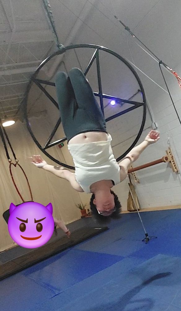 Nonbinary Asian hanging by their knees on an aerial pentagram. They're wearing glasses, a white tank top, and black leggings.