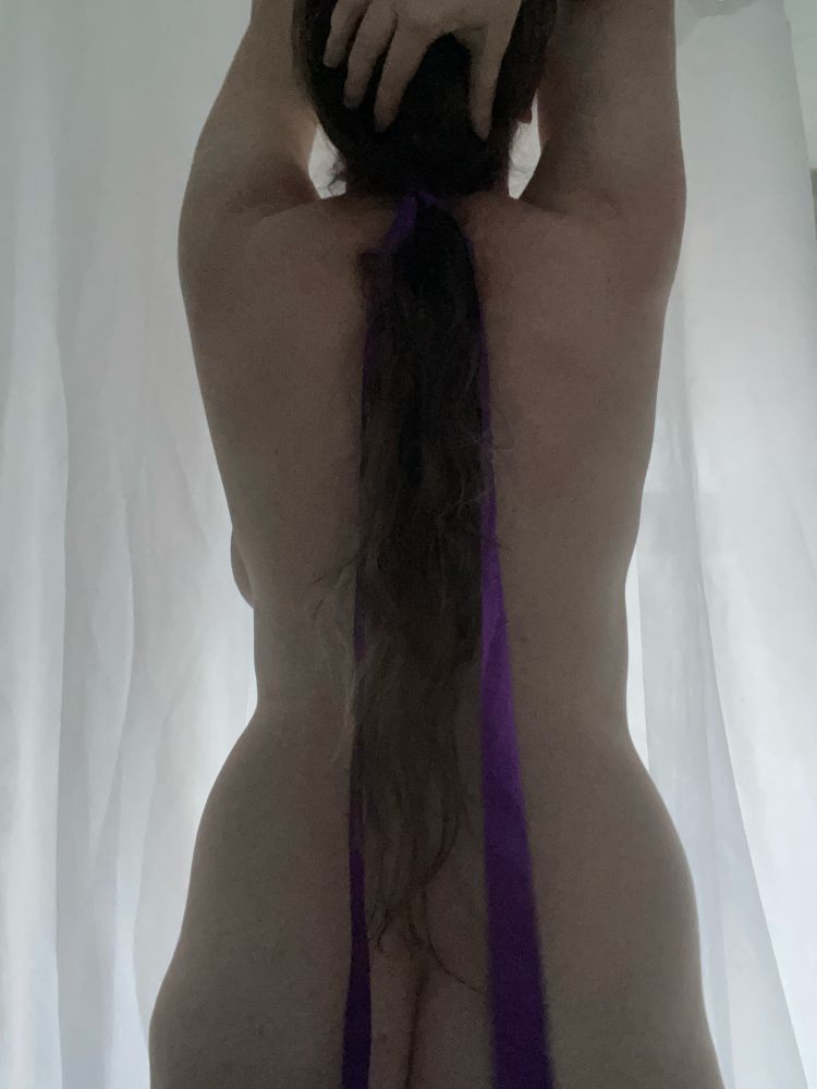 A white woman with her arms up and her back to the camera in front of a white background. She is naked and her long brown hair is tied with a purple ribbon.