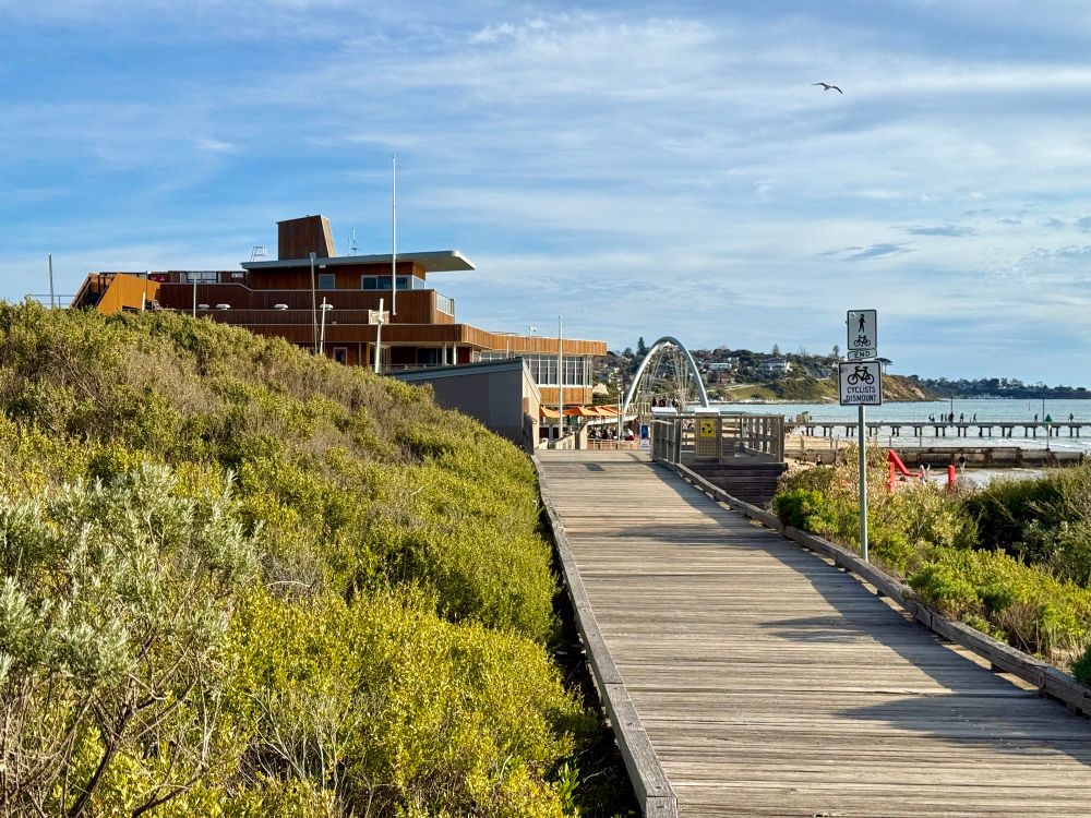 view along a timber boardwalk past low vegetation to a timber-clad building and arched bridge