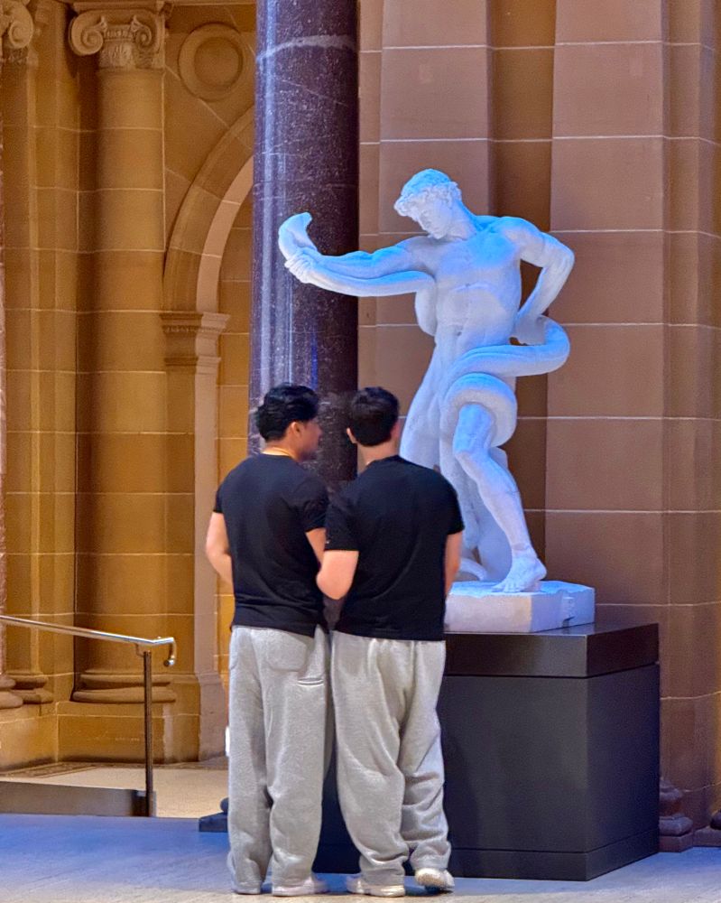 two identically dressed young men look at a homoerotic sculpture of a naked man writhing with a serpent