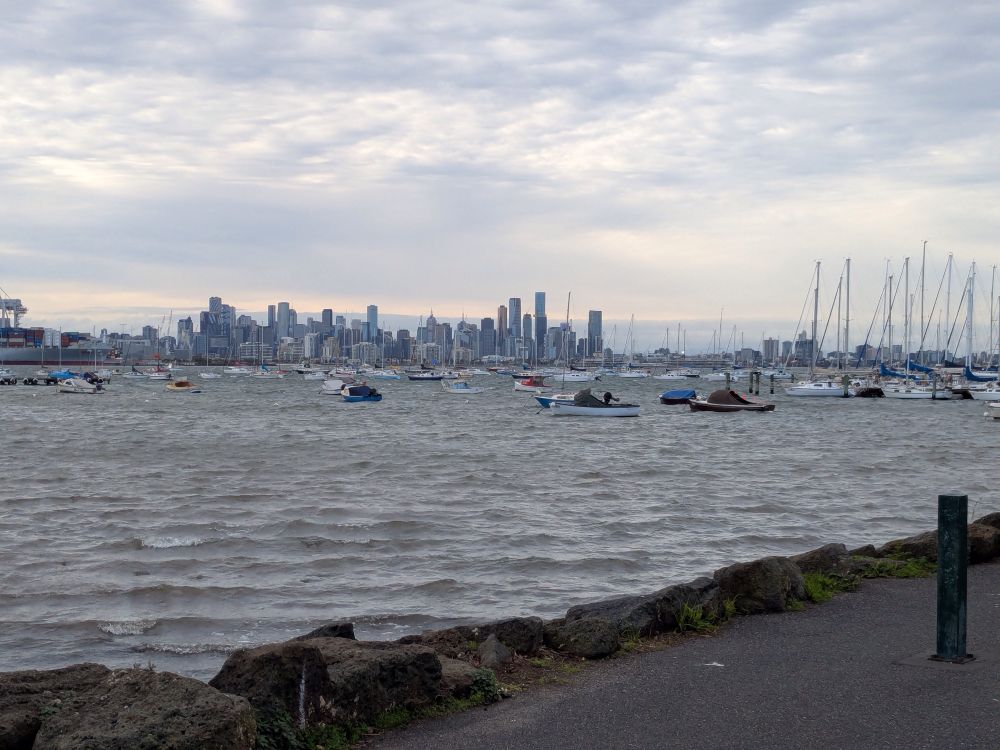 A wider angle shot with buildings and tall masts visible in the background against a sky that has strips of light and grey. 
The water is the middle ground laps against the brown strip of pathway. 