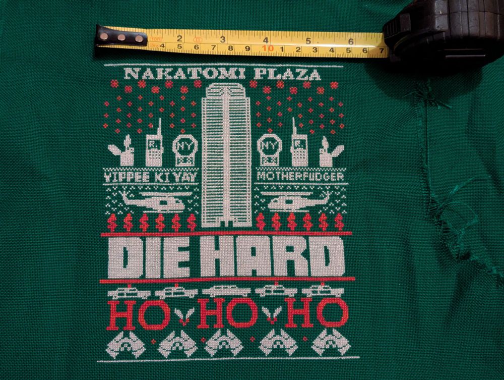 Green fabric with a white and red Die Hard design in the style of a Christmas jumper. I tweaked the wording as the recipient has a child so it says Yippee Ki Yay Motherfudger. My favourite bit is Nakatomi Plaza. There is also a tape measure to demonstrate that the whole thing is only 15cm wide.