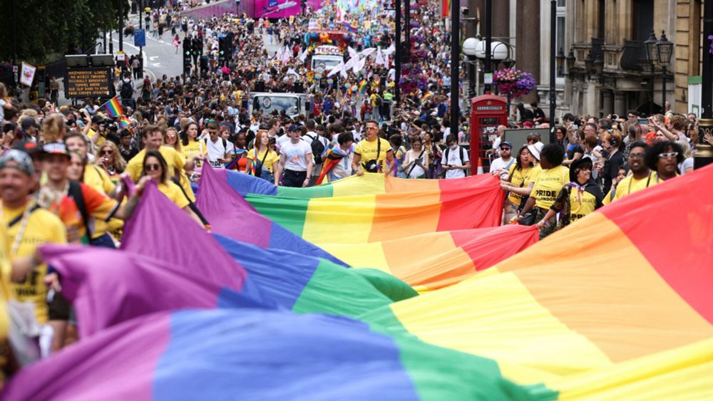 A scene from London's Pride Parade, featuring a large crowd holding and walking beneath a massive, colourful rainbow Pride flag as they march down a busy street.
