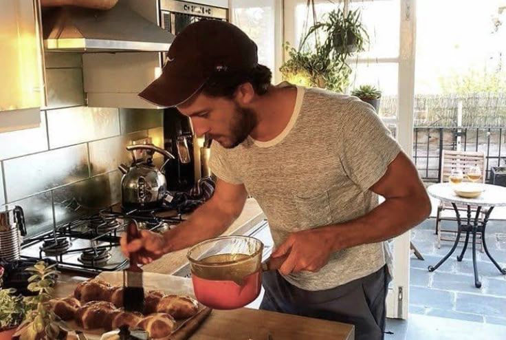 The actor Jonathan Bailey cooking and glazing bread rolls in a sunny kitchen.