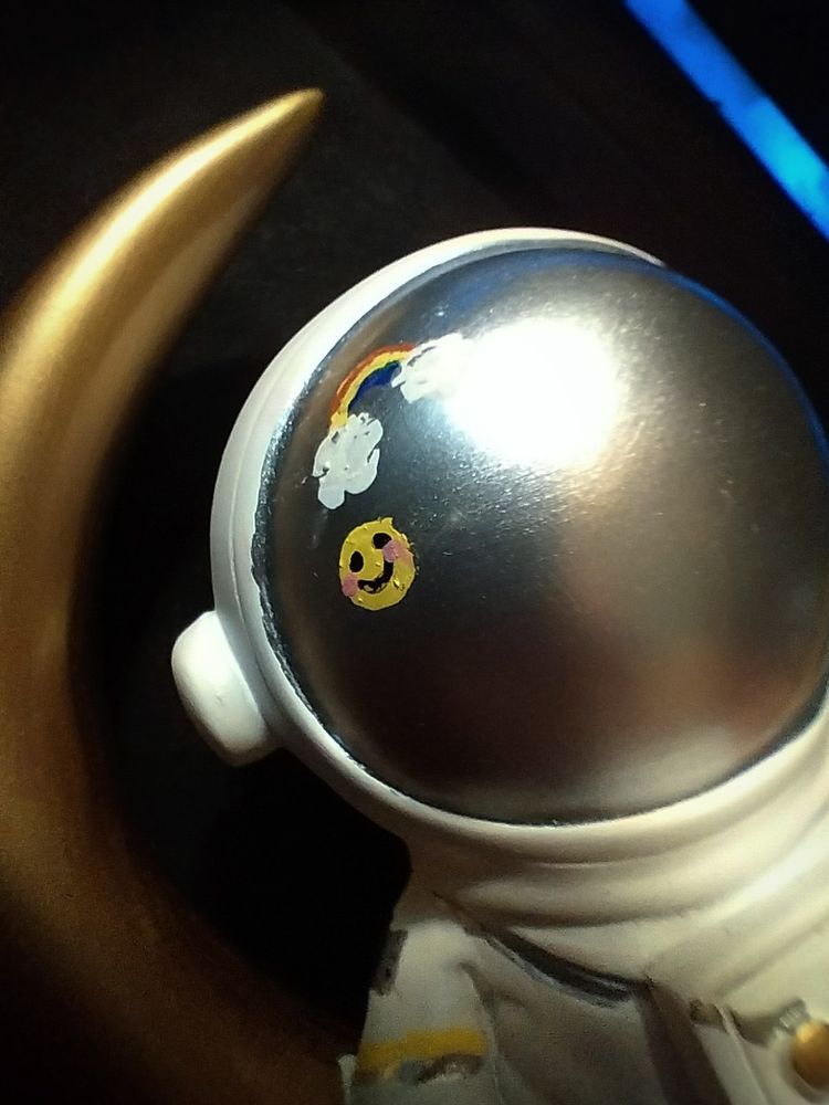 Astronaut / now Tessa Eliot from murder drones sitting in a crescent moon & laying down. A small smiley blushing sticker face and rainbow with clouds is painted on the counter of the helmet.
