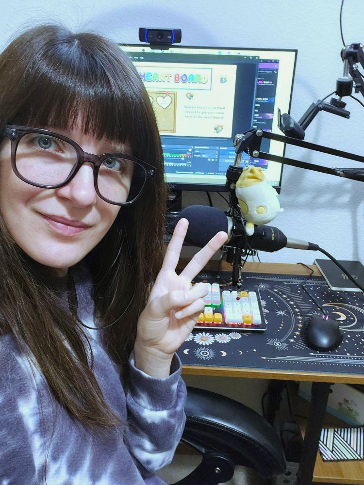 A photo of a person. Bee, the streamer, is sitting in front of their desk, smiling and making a peace sign. They are wearing large framed glasses and a purple tie-dye sweater. In the background, on top of their desk, is their monitor with a webcam on top, and streaming software pulled up. Also on their desk is their microphone, mouse, keyboard, and mouse mat.