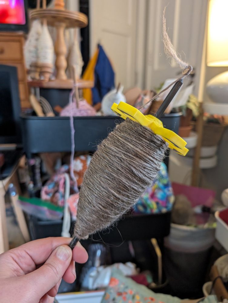 a yellow modular top whorl drop spindle from akerworks is FULL of heathered grey single spun yarn. the spindle is held by a white person's left hand, and the background is a blurry and chaotic assortment of spinning accoutrements 