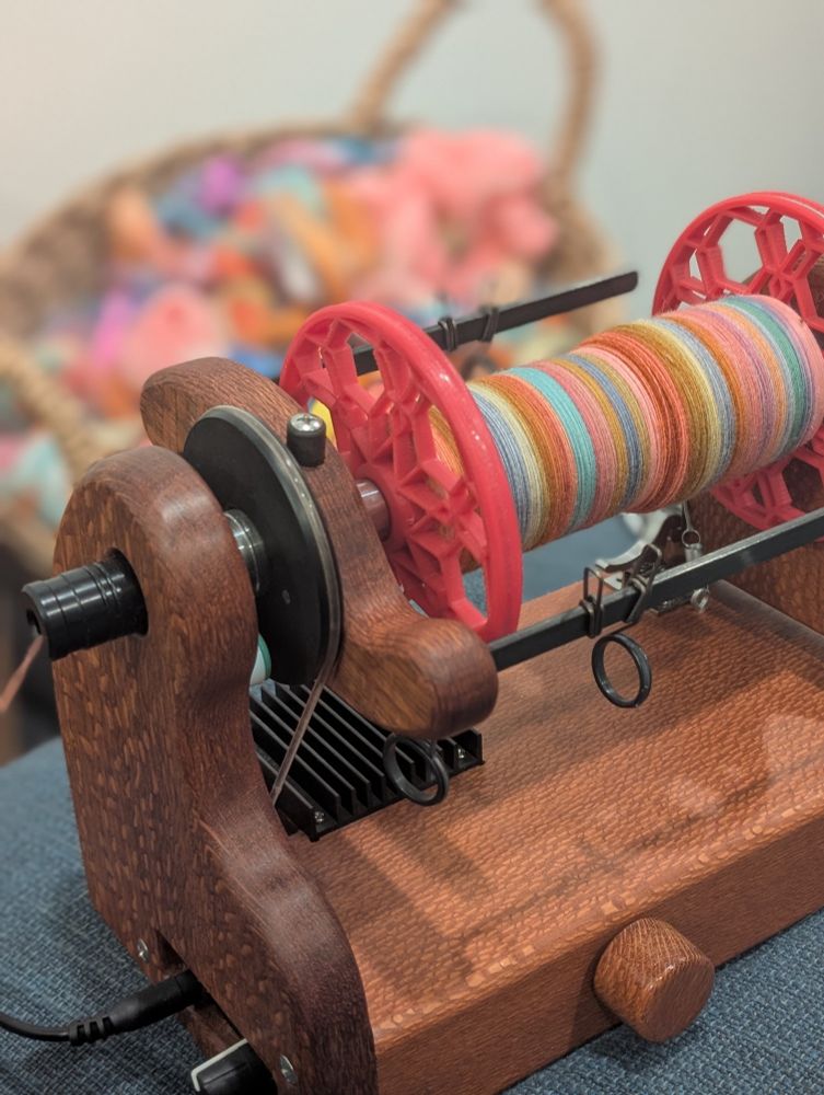 a hot pink bobbin is setup on a lacewood electric spinning wheel, and the bobbin is about a third full of a pink-dominated combo spin of very fine singles. in the background is a blurry basket of colorful, mostly pink, fluffy wool