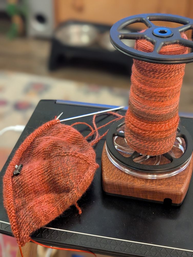 Brenna's latest work in progress, a hat knit with the Musselburgh pattern, sits atop a closed laptop. the project is connected to a bobbin of yarn, in reddish/pinkish and brown hues, which Brenna was clearly too lazy to wind into a center pull ball. She will unwind from the bobbin as she knits.