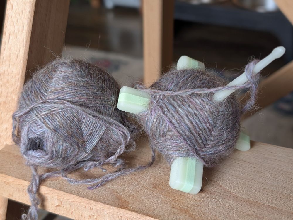 close up of an in-progress two-ply yarn on a glow in the dark 3d printed spindle. the yarn being plied comes from a center pull ball made with a cross arm spindle, and the yarn is a heathered grey-purple
