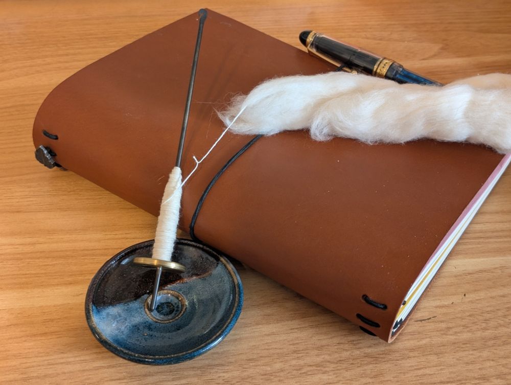 a tahkli spindle, traditionally used to spin cotton and other short stapled fibers, rests in a small blue and black ceramic spinning dish and is leaning against a warm brown leather journal. the fine, slubby cotton single being spun on the support spindle is connected to a small piece of white cotton fluff. it is nowhere near being full -- this is a project that will occupy a maker for quite some time.