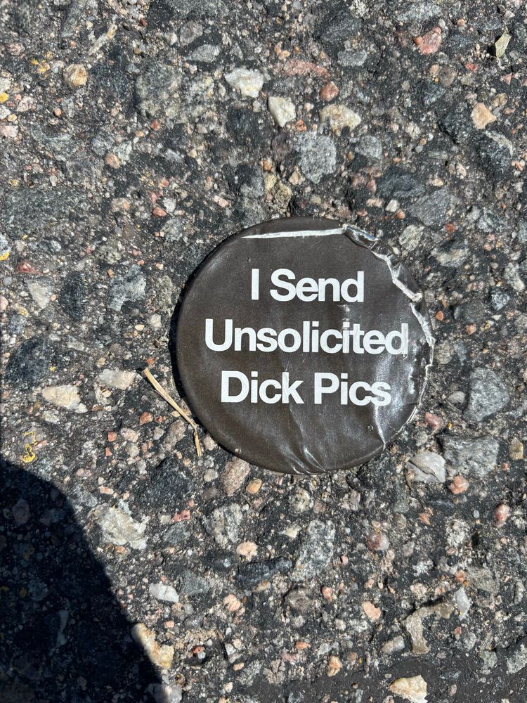 A button (white text on black background) lying on pavement. It reads “I Send Unsolicited Dick Pics”