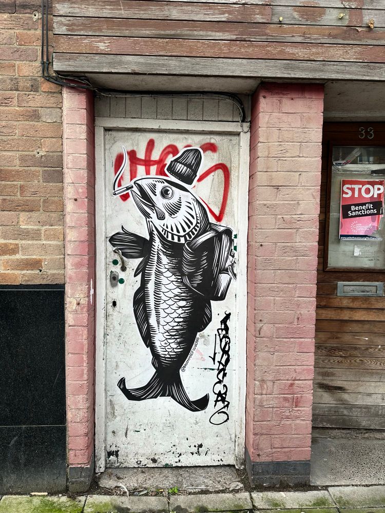 A piece of street art depicting a fish with a hat and rucksack, on an old door with a Stop Benefits Sanctions poster to the right. 
