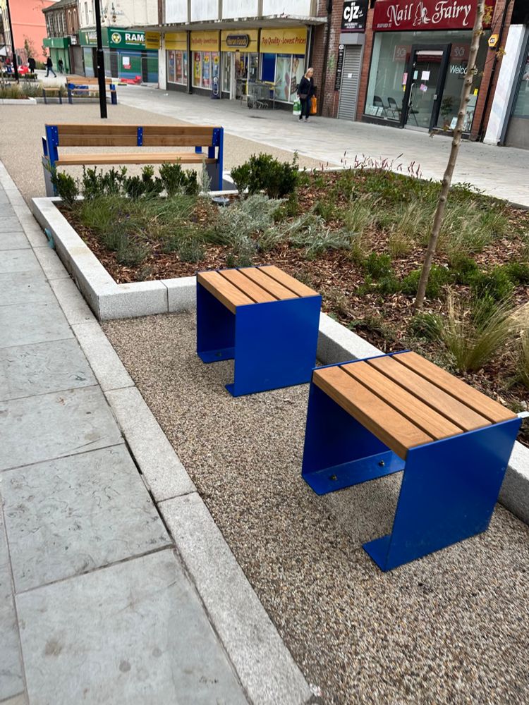 New seating, blue legs and wooden slatted seats, with combinations of single seats and benches placed around planters, on Bedford St, North Shields 