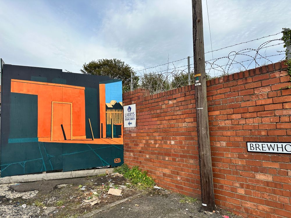 Anthony Downie’s mural in context, with rubble, rubbish and weeds, a Larry’s Fishcakes sign, barbed wire, a telegraph pole, a brick wall, and half a sign saying Brewhouse Bank. 