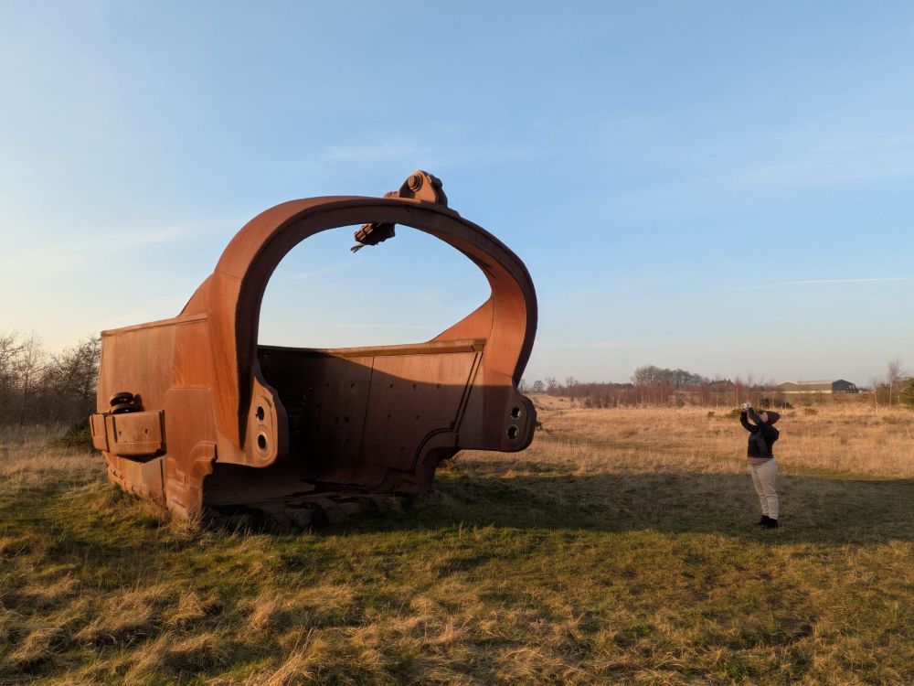 Orange iron giant bucket scoop on grassland, woman stood taking photo of it is tiny in comparison