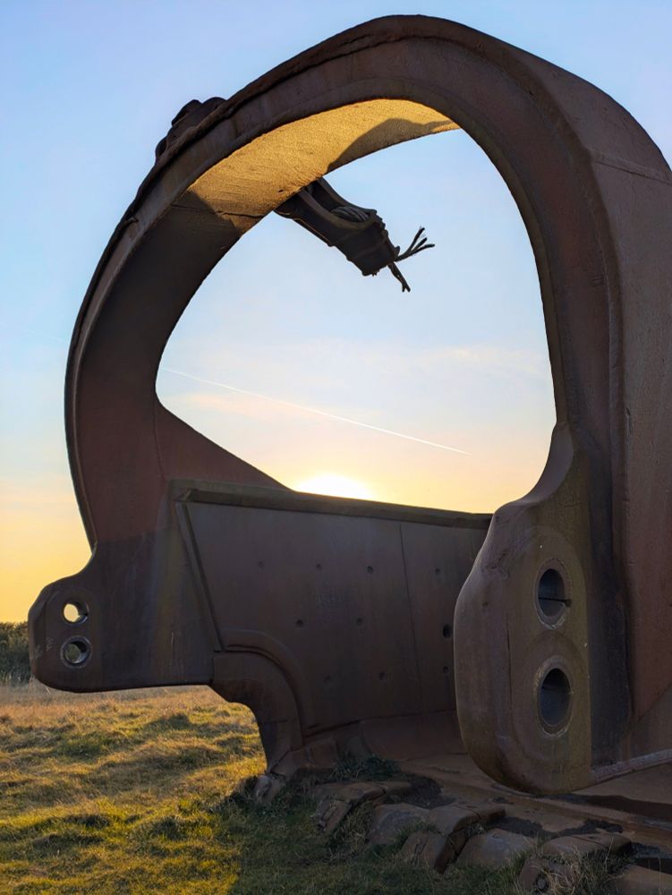 Orange iron giant bucket scoop on grassland, sunset seen behind 