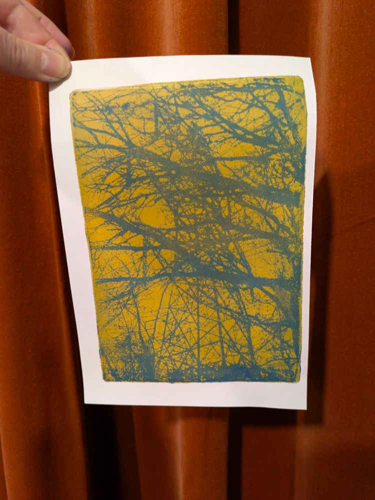Hand holding up a print of tree branches with electricity pylon tower behind. The print is ochre and blue, and is being held in front of orange velvet curtains 