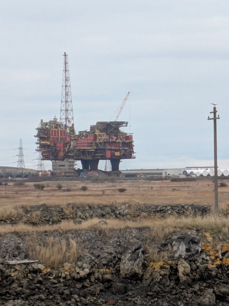 The Brent Charlie oil rig with fields of straw-like grass and rocks in foreground.