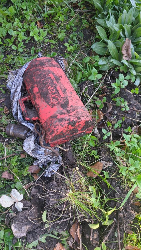 A red plastic crayola box covered in mud, resting on muddy ground and surrounded by grass and plants. 