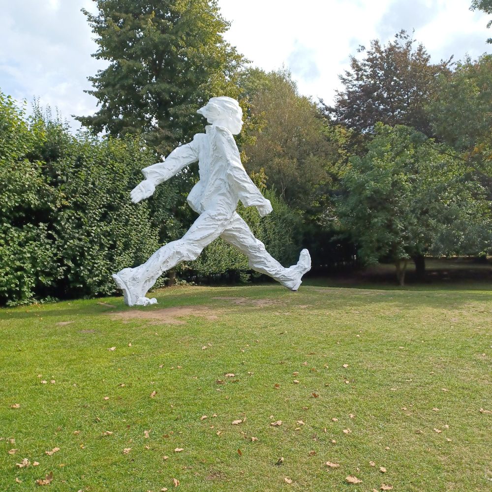 Eine zielstrebig gehende, riesige weiße Skulptur vor grünen Büschen. Sie erinnert ein wenig an einen überlebensgroßen Nils Holgersson.