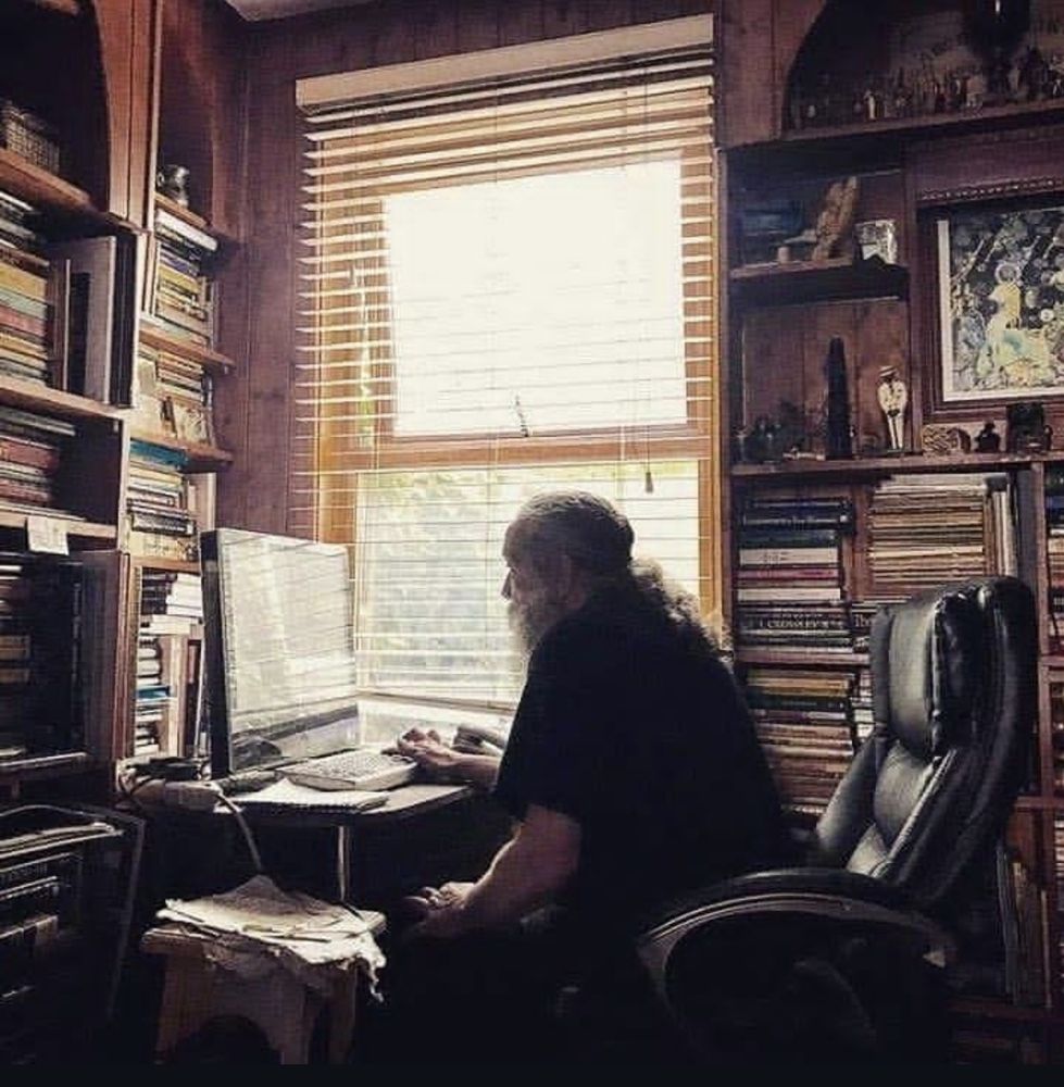 Alan Moore em frente ao computador no sei escritório, de barba comprida grisalha e cabelo preso, com prateleiras repletas de livros. Numa delas, atrás dele, à direita da foto, uma discreta imagem do Zé Pilintra entre outros itens de decoração 