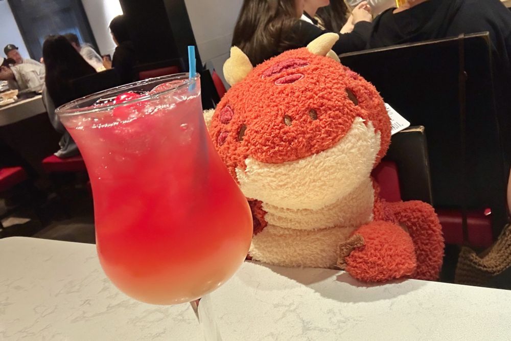 a red plush dragon created by fluffnest with a red drink in front of him. he is in a restaurant. 