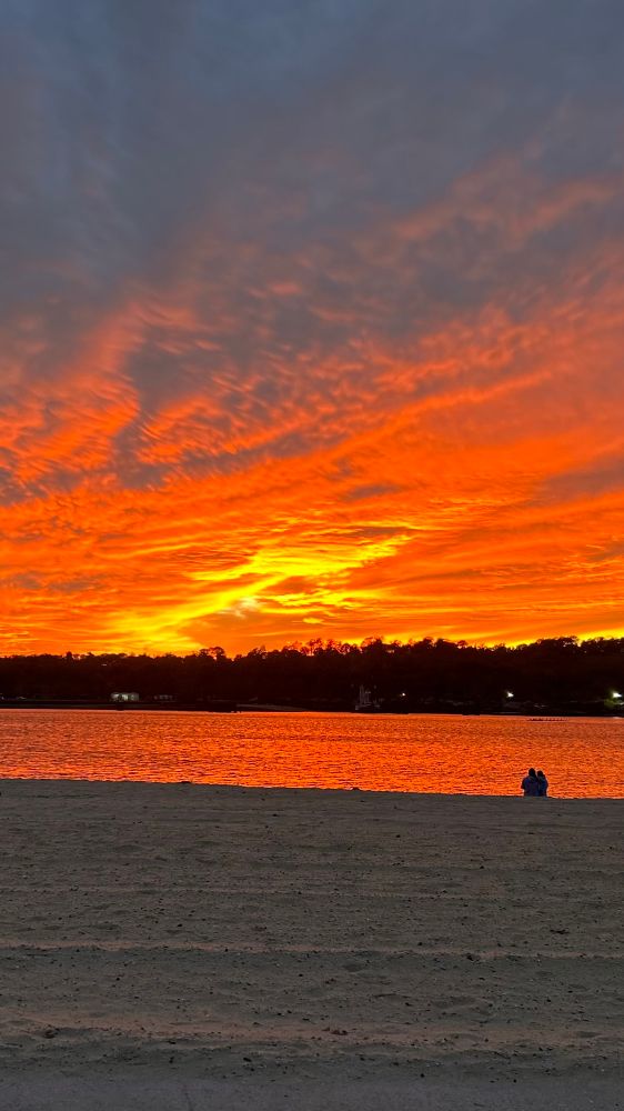 The horizon is lined with a low silhouette of trees and faint lakeside structures, but they recede into near-darkness against the brilliance above. The body of water stretches across the mid-frame, its surface turned to liquid fire as it reflects the sunset’s deep oranges and molten golds. Every small wave glimmers as though lit from within.

Above this glowing band, the sky erupts in sweeping, textured clouds—broad strokes and feathered patterns radiating outward as though the entire heavens have been brushed with flame. The light intensifies toward the horizon, where the brightest core of the sunset burns in a fierce concentration of yellow and incandescent orange. As the eye travels upward, the color transitions gradually into muted rose, then into a cooling slate-blue that signals approaching nightfall.

The sandy foreground is dim and undisturbed, its pale tones now subdued in the fading light. Near the waterline, two small figures sit closely together, wrapped or bundled against the evening air. Their presence adds a quiet human scale to the grandeur of the scene, emphasizing the vastness of the sky above them and the peaceful stillness of the beach.