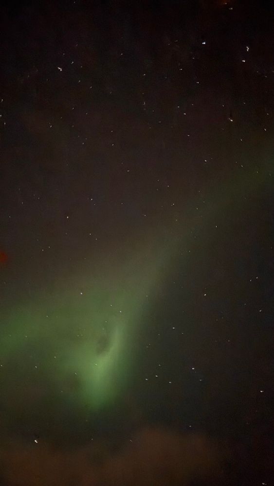 A quiet stretch of night sky opens above, sprinkled with sharp, scattered stars that glimmer against a deep, velvety darkness. Through that stillness, a soft green arc of aurora drifts upward—subtle, smooth, and faintly luminous, like a breath of light rising from the earth itself.

Near the center, the aurora thickens into a gentle swirl, its glow folding around a darker patch of cloud, giving the impression of a slow-turning celestial eddy. Wisps of cloud hover along the edges, catching just enough light to form hazy borders around the glow