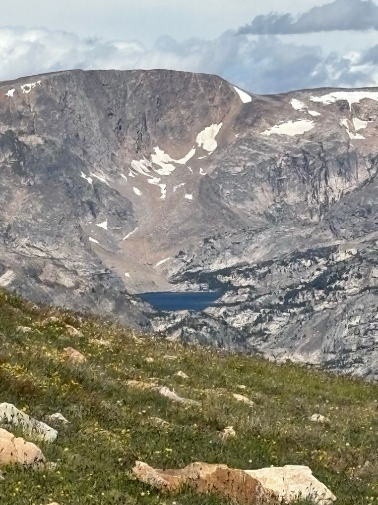 The image captures a rugged and dramatic view of the Rocky Mountains. In the foreground, a grassy alpine meadow is sprinkled with small yellow wildflowers and dotted with pale, weathered boulders. Beyond the meadow, the terrain drops sharply into a vast, rocky basin. The jagged granite slopes rise steeply on all sides, streaked with patches of lingering snow clinging to crevices and sheltered ledges. Near the center of the scene, a deep blue alpine lake sits nestled far below, its still waters contrasting vividly with the surrounding gray and tan stone. In the background, a towering mountain ridge dominates the horizon, its summit bare and windswept under a partly cloudy sky, with soft, billowing clouds adding depth to the view. The overall feel is one of remote wilderness and raw, untamed beauty.