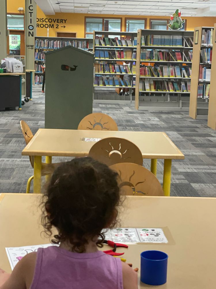 Ellie sitting at a table coloring at the library, with bookshelves as far as the eye can see.