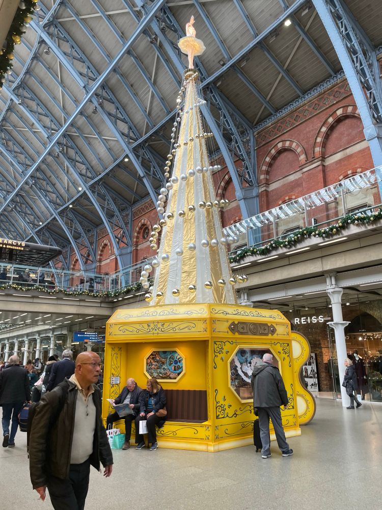 A bright yellow helter-skelter type structure with a spiral of baubles, in a high ceiling mall.