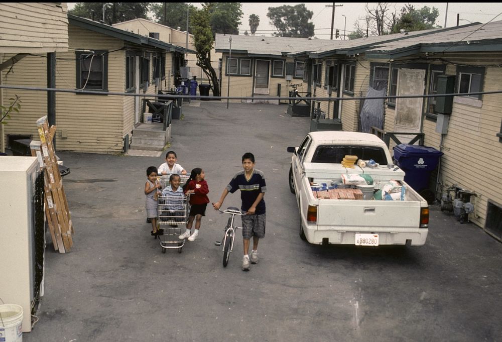 A paved courtyard in Los Angeles. Danny, Inside the shopping cart, Angel white shirt, Letty, red dress, Kevin, small boy. Alley w. of Wall St., north of 41st St., Los Angeles, 2002
