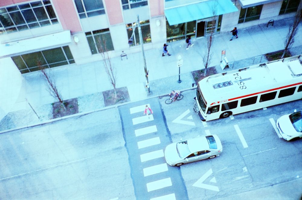 looking down from above at a crosswalk with one person crossing and one bike, one bus, and two cars waiting at the crosswalk