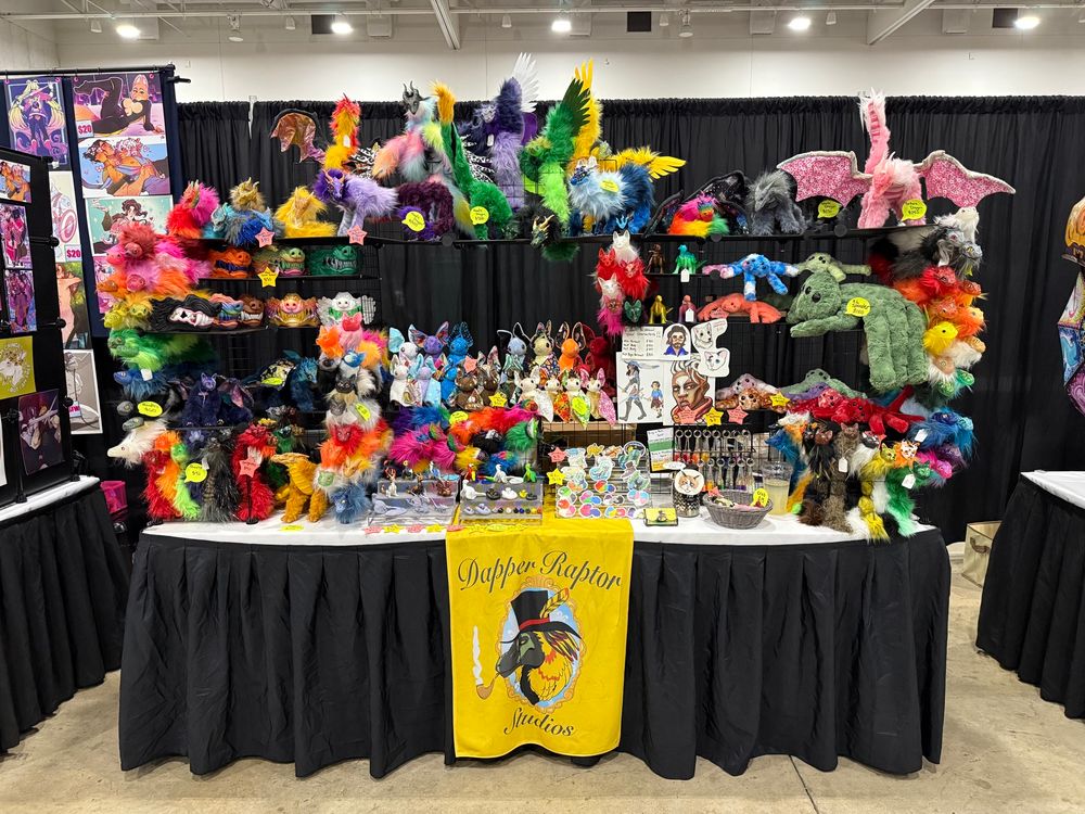 Booth set up with banner that says Dapper Raptor Studios and various art dolls of dragons and other creatures all around. Also plushie bats and spiders. 