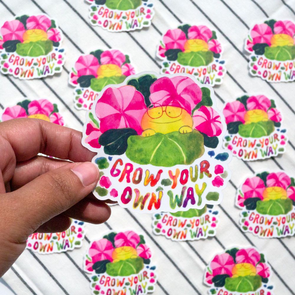 I'm holding up a sticker with an illustration of a round frog between flowers and the text says "grow your own way"