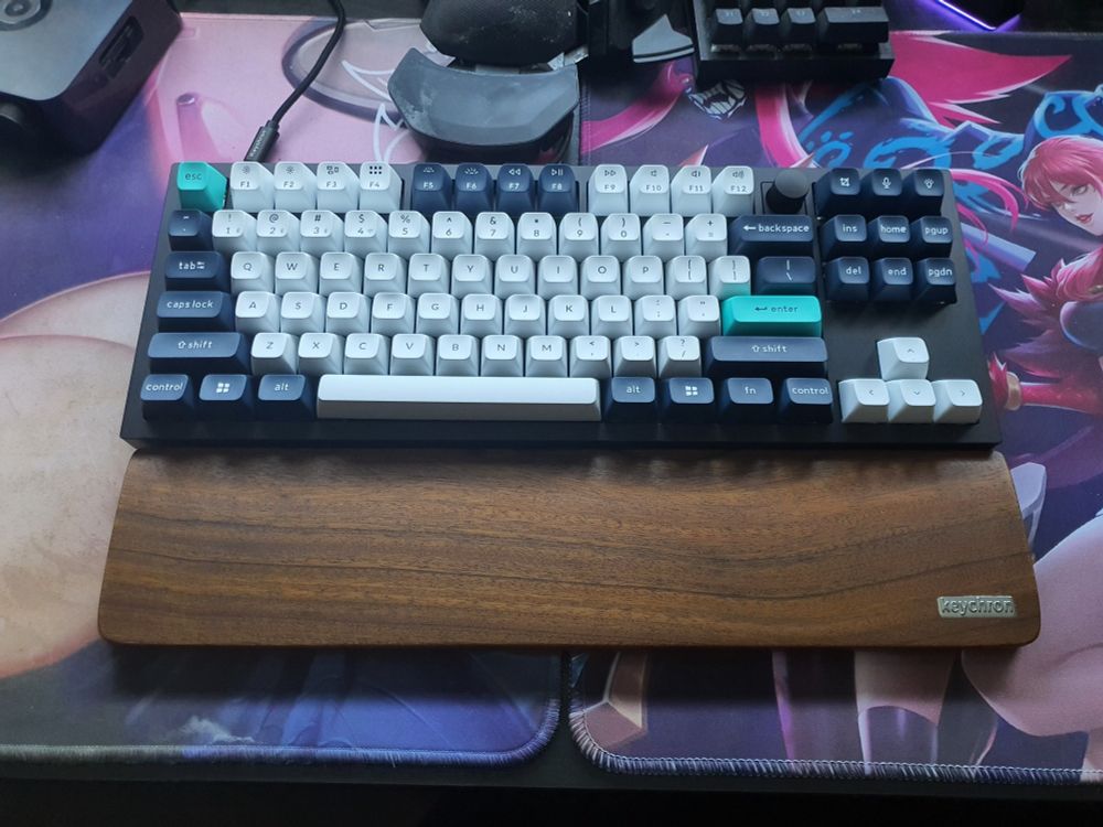 A TKL keyboard with a wooden wrist rest, sitting between two colorful mouse mats. the keyboard is a Keychron Q3 Max