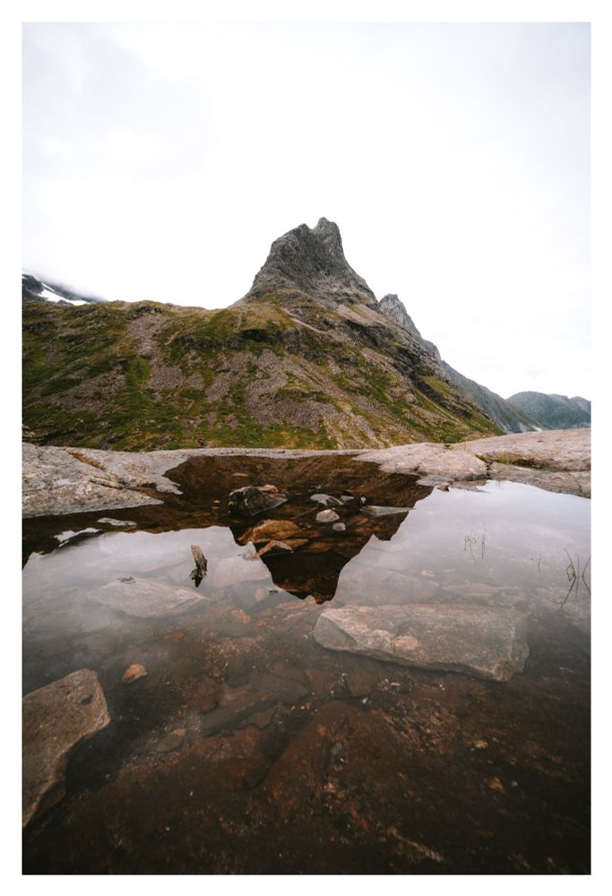 A rocky mountain peak rises above green and rocky slopes, reflected in a shallow pool of water with stones visible beneath the surface under an overcast sky.