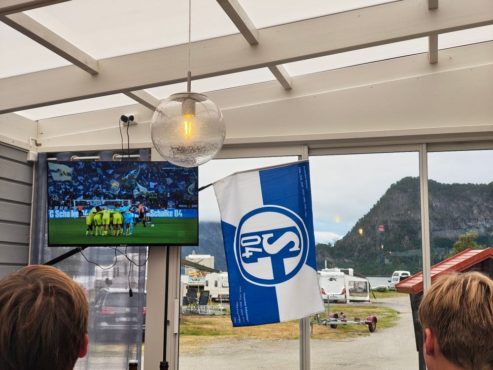 Foto von einem Fernseher, einer Schalke Fahne im einen Glashaus und außen ein Campingplatz und Berge im Hintergrund.